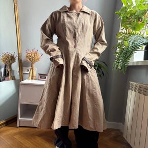 May include: A long-sleeved, knee-length beige coat featuring a pointed collar, a front button closure, and dark brown decorative straps with buckles on the cuffs. The coat has a flared, A-line silhouette with visible vertical seam detailing.
