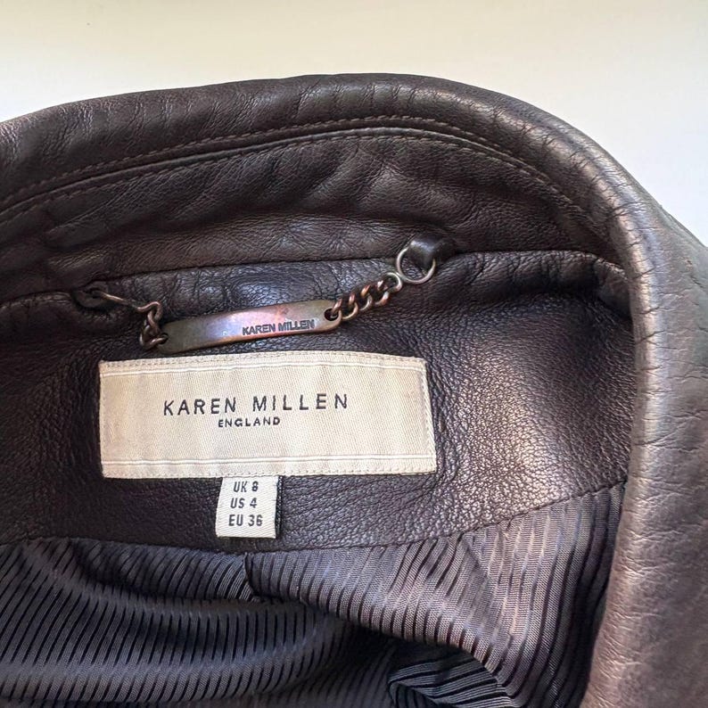 May include: Close-up of a dark brown leather jacket interior. The jacket features a label that reads "KAREN MILLEN ENGLAND". The jacket has a chain detail and a size tag showing UK 6, US 4, and EU 36.