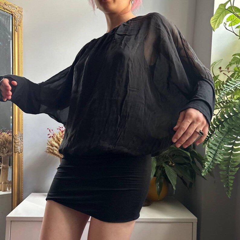 May include: A black long-sleeve blouse featuring a sheer, loose-fitting bodice with a round neckline and wide batwing sleeves. The garment has a fitted, solid black hemline that sits at the waist, creating a blouson effect with a draped silhouette.