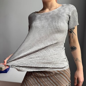 Puede incluir: Una camiseta gris de manga corta con un patrón texturizado en forma de rejilla. La tela parece ligeramente transparente y tiene un aspecto arrugado único. La camiseta tiene un cuello redondo y una silueta ajustada.