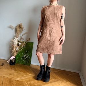 Puede incluir: Vestido de ante marrón sin mangas, hasta la rodilla, con una silueta sencilla en forma de A. El vestido presenta costuras verticales y diagonales. La persona lleva botas negras con plataforma. El vestido mide unos 90 cm de largo.