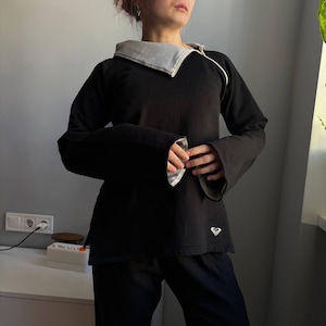 May include: Black long-sleeved top with a contrasting white and grey collar and cuffs. The top features a unique design with flared sleeves and a small logo on the lower left. The collar has a textured pattern.
