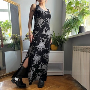 May include: A sleeveless black maxi dress featuring a white floral pattern, a scoop neckline, and a high side slit. The dress is paired with black mid-calf boots with metallic hardware details, creating a cohesive look with a simple silhouette.