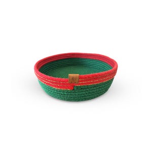 May include: A green and red rope basket with a gold trim. The basket is made of thick, woven rope and has a round shape.