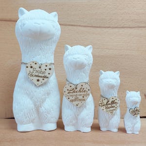 Könnte beinhalten: Vier weiße Alpaka-Figuren aus Keramik in verschiedenen Größen. Jedes Alpaka hat ein herzförmiges Holzschild um den Hals mit deutschem Text. Das größte Alpaka hat "Frohe Weihnachten" auf seinem Schild. Die anderen Schilder haben verschiedene deutsche Sätze.