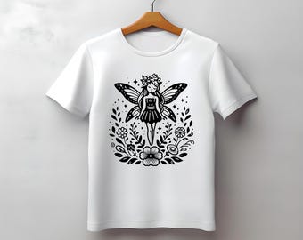 Puede incluir: Camiseta blanca con una ilustración en blanco y negro de un hada. El diseño incluye un hada con alas de mariposa, una corona de flores y un vestido, rodeada de flores y hojas. La camiseta está en una percha de madera.