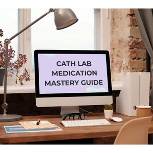Puede incluir: Una pantalla de ordenador muestra el texto "CATH LAB MEDICATION MASTERY GUIDE". El ordenador está sobre un escritorio de madera con teclado, ratón y gafas. También se ven una lámpara de escritorio y una planta en maceta.
