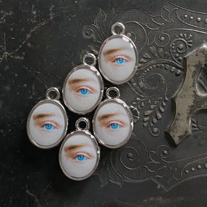 Puede incluir: Cinco abalorios ovalados de plata con un ojo azul y una ceja impresos en cada uno. Los abalorios tienen un bucle en la parte superior para sujetarlos a un collar o pulsera.