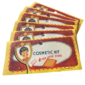 May include: A stack of vintage cosmetic kits with a yellow background and a red and white oval design. Each kit features a portrait of a young person holding a mirror and the text "Cosmetic Kit for Little Stars."