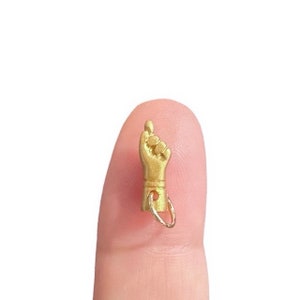 May include: A small, gold-colored charm in the shape of a hand with the index finger pointing up. The charm is attached to a small ring. The charm is shown on a fingertip.