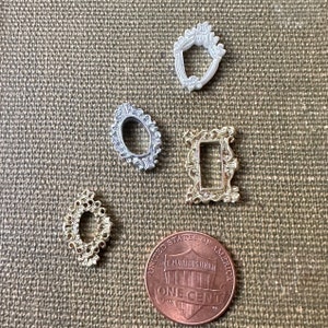 May include: Four miniature ornate picture frames in silver and gold tones. The frames are arranged on a brown fabric surface with a US penny for scale.