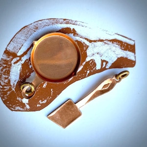 May include: A copper-colored miniature frying pan with a small spatula. The pan has a round cooking surface and a handle with a loop. The spatula has a rectangular head and a long handle. The items are on a brown and white patterned surface.