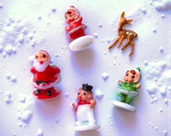 May include: A collection of miniature Christmas figurines, including Santa Claus, a snowman, an elf, and a reindeer, are arranged on a snowy surface. The figurines are brightly colored with red, white, green, and brown hues, evoking a festive holiday theme.