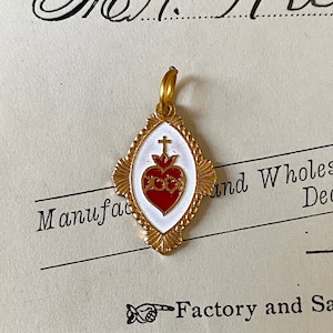 May include: A gold-colored pendant with a white enamel background and a red enamel heart with a cross on top. The heart has a gold border and three gold circles around it. The pendant is in the shape of a diamond.