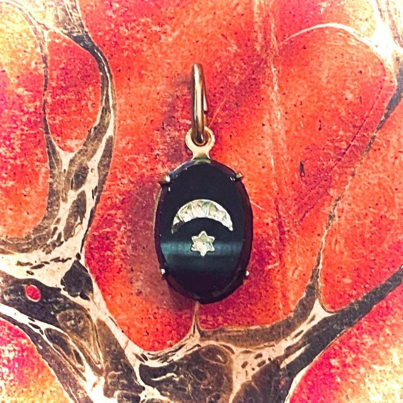 May include: An oval-shaped pendant with a black background, featuring a crescent moon and a star design. The pendant has a gold-colored bail and is set against a red and orange background.