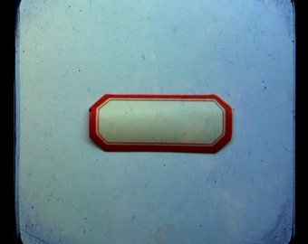 May include: A rectangular label with a red border and a white interior, centered on a light blue background. The label's shape is a rounded rectangle with eight sides. The background has a slightly textured appearance.