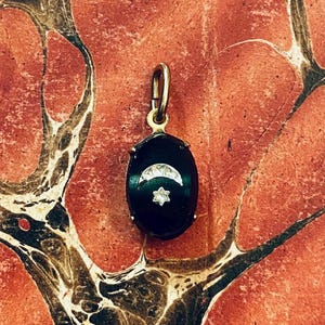May include: A gold-toned pendant with an oval, black onyx center. The onyx features a crescent moon and a star design, both in a lighter color. The pendant is suspended from a gold bail.