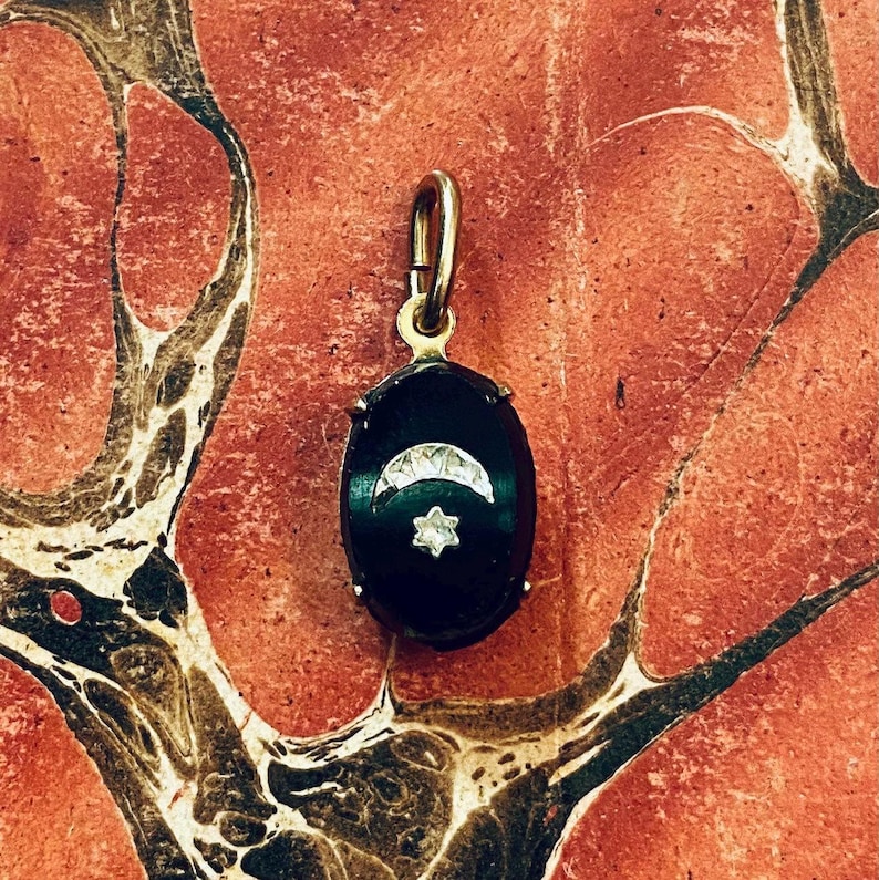 May include: A gold-toned pendant with an oval, black onyx center. The onyx features a crescent moon and a star design, both in a lighter color. The pendant is suspended from a gold bail.