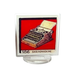 May include: A square, clear acrylic display stand holding a card with a vintage typewriter illustration. The card has a red background with a black border. The text "186 SKRIVEMASKINE" is at the bottom.