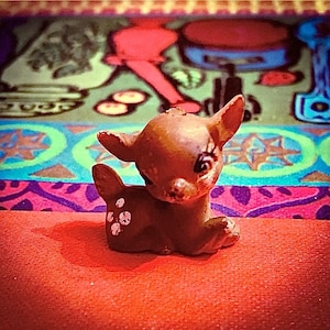 May include: A small, brown ceramic deer figurine with white spots on its back. The deer is sitting on a red surface, with a colorful, patterned background. The figurine has large eyes and is in a resting pose.