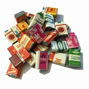 May include: A collection of miniature cigarette boxes in various colors, including red, white, green, and gold. Brands such as Lucky Strike, Camel, Winston, and Salem are visible. The boxes are arranged in a pile.