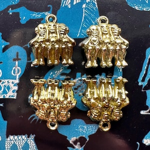 May include: Four gold-colored charms, each depicting a group of three figures. The charms have a loop at the top for hanging. The background is a blue fabric with a repeating pattern of white and black shapes.