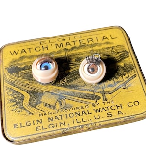 May include: Two antique watch eye charms with detailed irises, one blue and one brown, set on a cream-colored base with metal eyelashes. The charms rest on a vintage yellow Elgin Watch Material tin, with text reading "ELGIN WATCH MATERIAL".