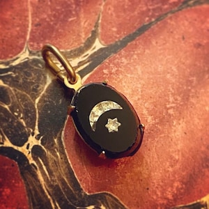 May include: A gold pendant with a black oval stone. The pendant features a crescent moon and a star, both embellished with small, sparkling stones. The pendant is suspended from a gold loop.