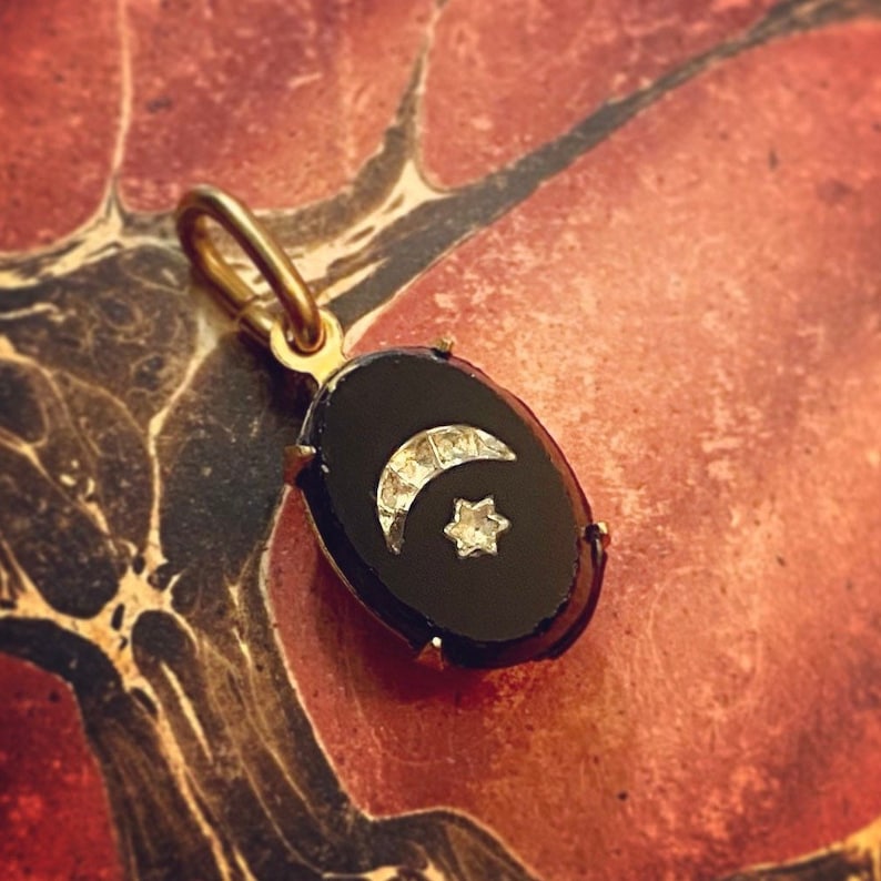 May include: A gold pendant with a black oval stone. The pendant features a crescent moon and a star, both embellished with small, sparkling stones. The pendant is suspended from a gold loop.