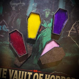 May include: Four miniature coffin-shaped decorations in various colors: yellow, purple, red, and pink. The coffins have dark brown frames and are set against a vintage horror-themed background. The coffins are open or partially open, revealing the colorful interiors.