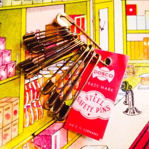 May include: A bundle of gold-colored steel safety pins with a red tag that reads "DOSCO STEEL SAFETY PINS MADE IN GERMANY". The pins are arranged in a cluster, with the tag attached. The background is a colorful illustration of a store.