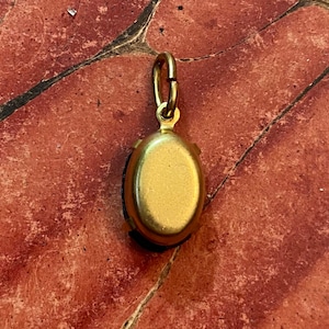 May include: A gold-colored pendant with an oval, dark-colored stone. The pendant has a small loop at the top for attaching to a chain. The pendant is set against a reddish-brown surface.