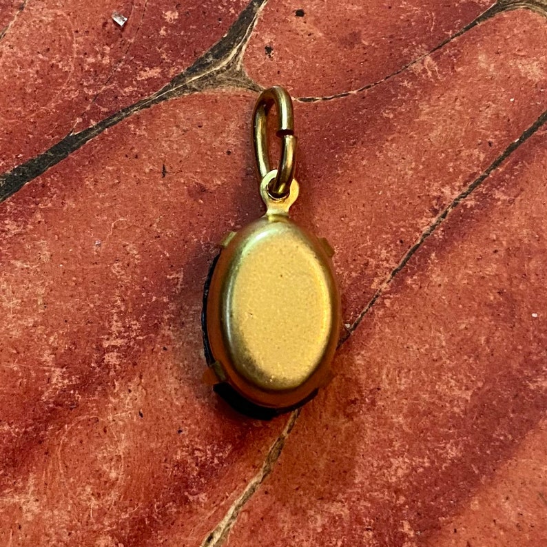 May include: A gold-colored pendant with an oval, dark-colored stone. The pendant has a small loop at the top for attaching to a chain. The pendant is set against a reddish-brown surface.