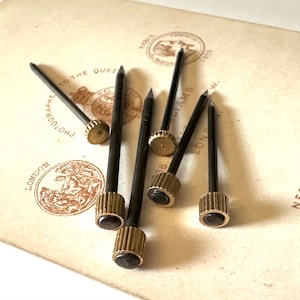 May include: Six antique brass tacks with black metal shafts. The tacks have a decorative, fluted design on the top. The tacks are arranged on a piece of paper with old stamps.