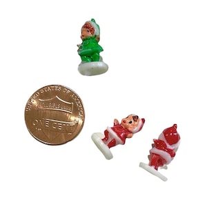 May include: Three vintage Christmas figurines. One is green and wearing a red hat, one is red and white and wearing a red hat, and one is red and white and wearing a blue hat. The figurines are standing on white bases.
