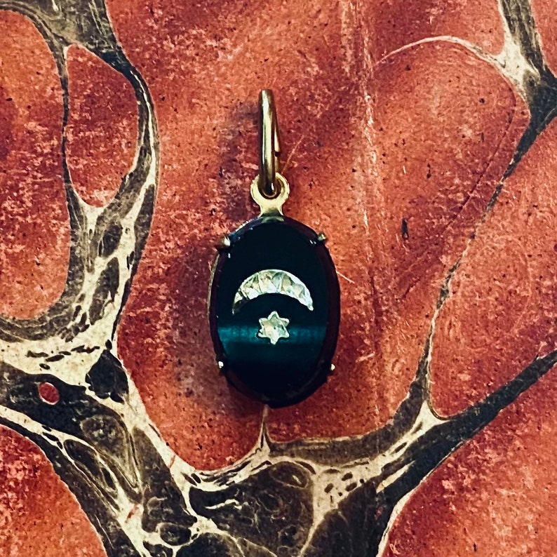May include: An oval pendant with a dark, possibly onyx, background. A crescent moon and a six-pointed star are set in the center. The pendant has a gold-colored bail and frame. The background is a marbled red and brown.