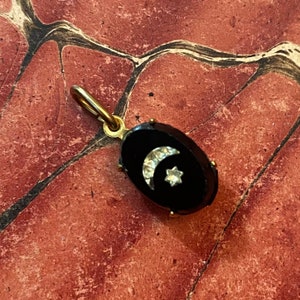 May include: An oval-shaped pendant with a black background, featuring a crescent moon and a star design. The moon and star are embellished with small, sparkling stones. The pendant has a gold-colored bail for attaching to a chain.