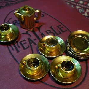 May include: A miniature gold tea set with a teapot, cups, and saucers. The set is arranged on a red velvet surface with the word "Tea-Table" printed on it.
