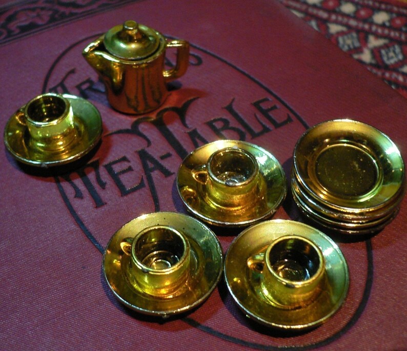 May include: A miniature gold tea set with a teapot, cups, and saucers. The set is arranged on a red velvet surface with the word "Tea-Table" printed on it.