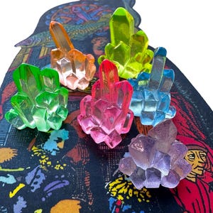May include: A set of six colorful crystal-shaped glass paperweights. The paperweights are in shades of green, pink, blue, purple, and yellow. They are arranged on a black and white patterned surface.