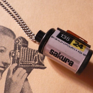 May include: A silver chain necklace with a silver and black film canister pendant. The canister is labeled "Sakura 135 24 Exposures 24.36mm ASA 100".