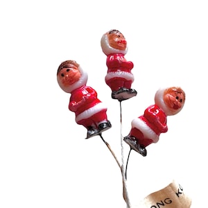 May include: Three small plastic figurines of Santa Claus wearing red and white outfits with white fur trim. The figurines are attached to wire stems and are arranged in a bouquet.