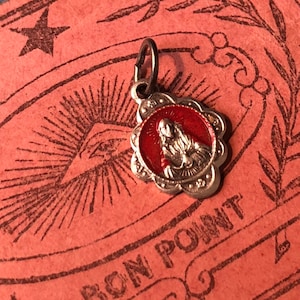 May include: A small, silver-toned pendant with a red center featuring a religious figure. The pendant has a scalloped edge and a small loop for a chain. The background is a pinkish-orange paper with a star and eye design.