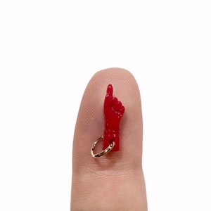 May include: A red plastic charm shaped like a hand with the index finger pointing up. The charm is attached to a gold ring.