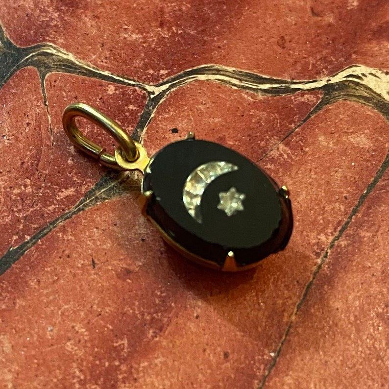 May include: A gold-colored pendant with an oval black onyx stone. The onyx features a crescent moon and star design made of small, sparkling stones. The pendant has a small loop at the top for a chain.