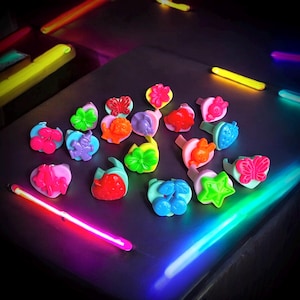 May include: A selection of brightly coloured plastic rings in various shapes, such as butterflies, flowers, and hearts. The rings are in shades of pink, green, orange, and blue. The image is lit with neon lights.