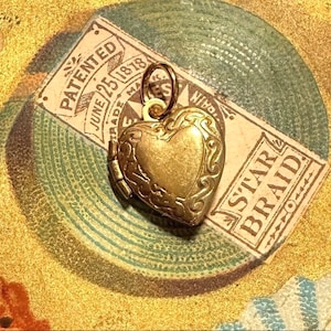 May include: A gold-toned heart-shaped locket charm with intricate details. The charm has a hinged opening and is attached to a gold-toned chain. The charm is sitting on a vintage-looking surface with text that reads "PATENTED JUNE 25 1878 TRADE MARK" and "STAR BRAID".