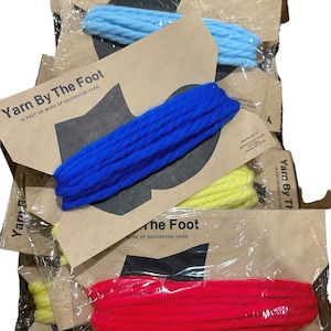 May include: Four skeins of yarn in different colors: blue, yellow, red, and light blue. Each skein is wrapped in plastic and labeled "Yarn By The Foot 15 feet or more of decorator yarn".