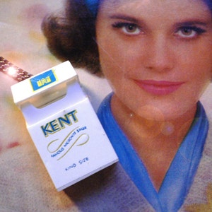 May include: A white and blue Kent King Size cigarette pack with gold lettering and a blue and gold logo. The pack is in front of a vintage advertisement featuring a woman with blue accents.