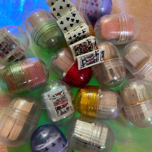 May include: A collection of clear plastic capsules filled with various playing cards. The capsules are arranged in a random pattern on a green surface. Some of the cards are visible through the clear plastic.